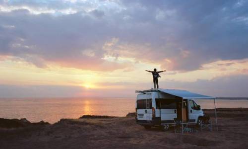 Young Caucasian woman  dancing  on roof of camper van on seaside  at sunset